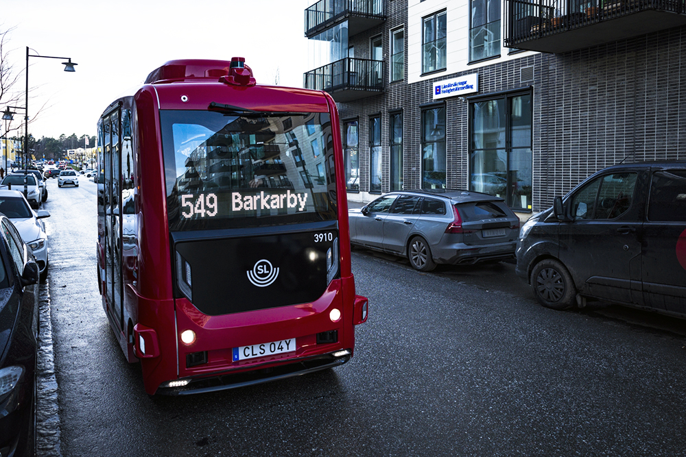 Självkörande buss kör på gata kantad av parkerade bilar.